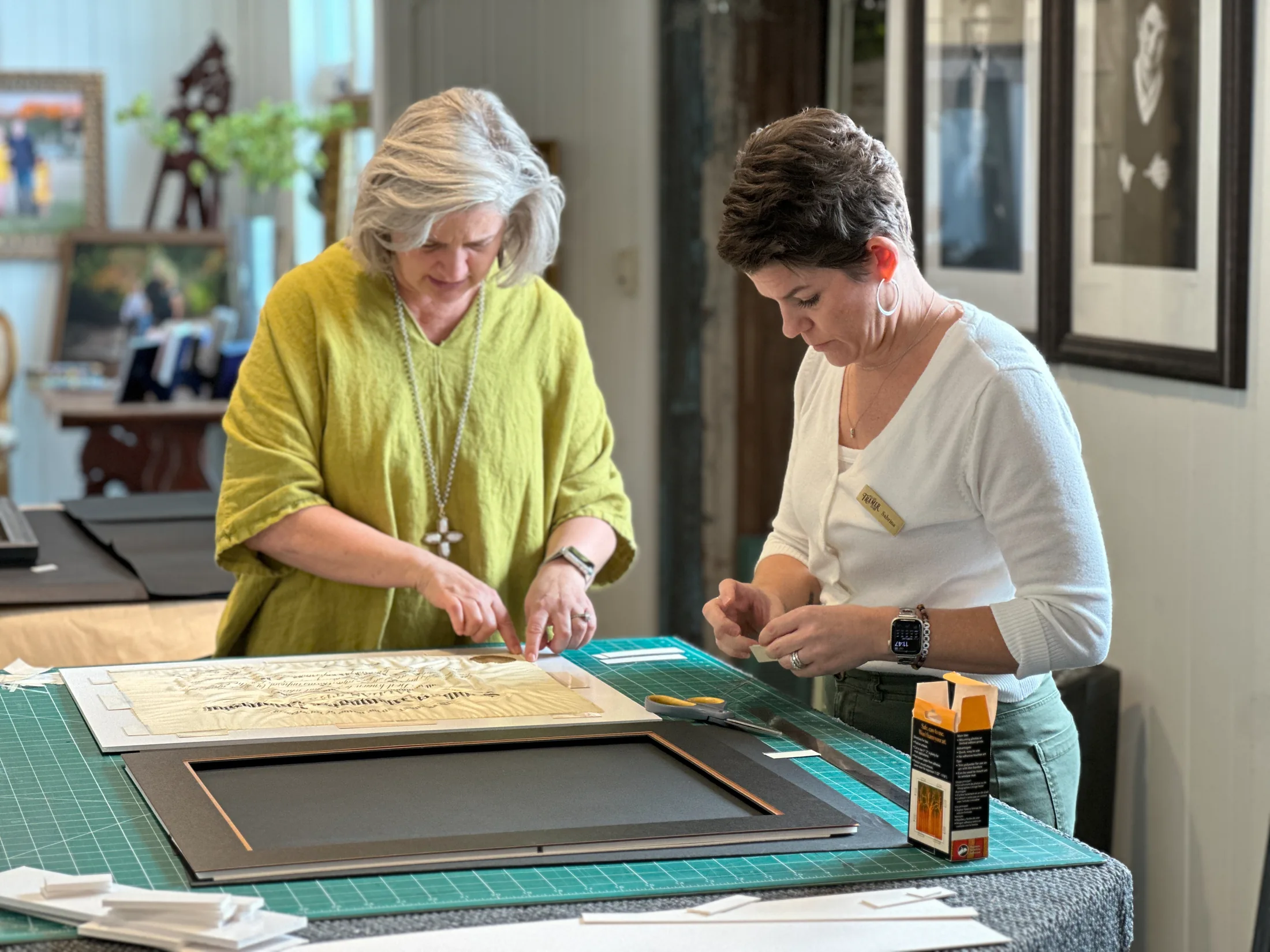 Two framers working together over artwork and matting at the Village Framer workbench